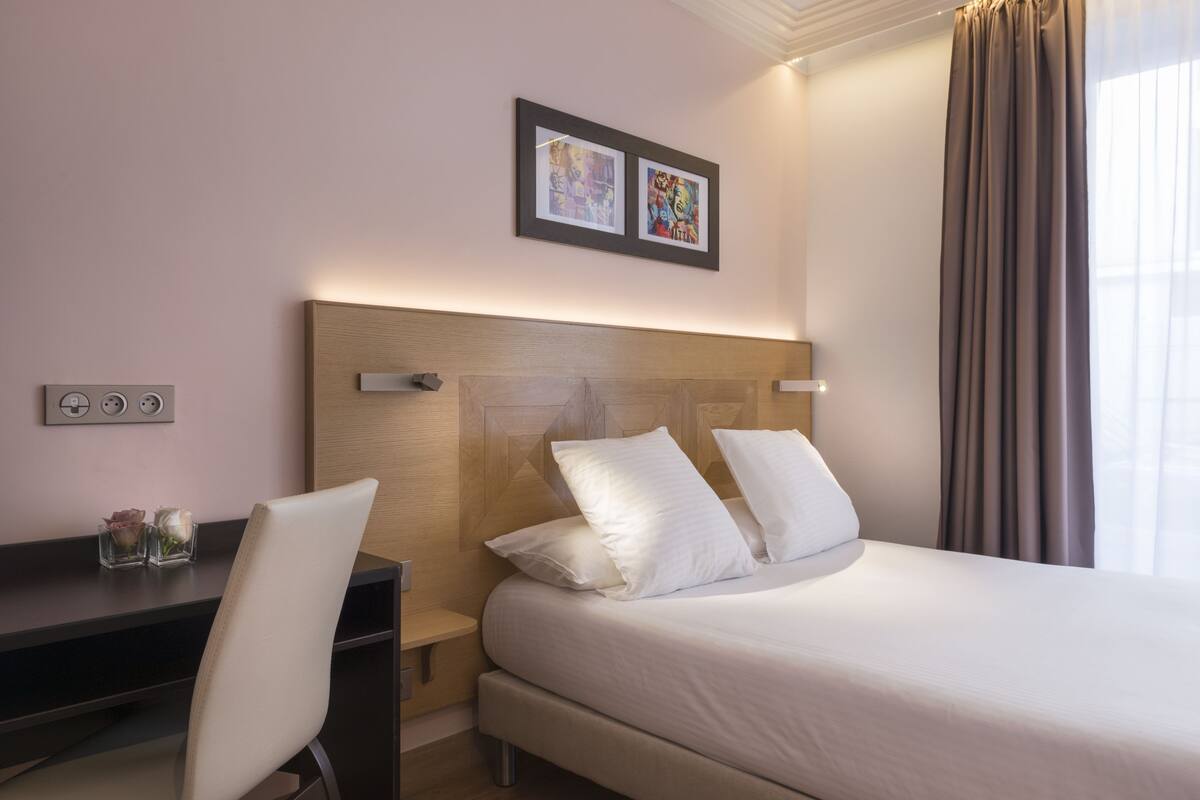 A hotel room with a bed, a desk, and a chair. There are two framed pictures on the wall above the bed.