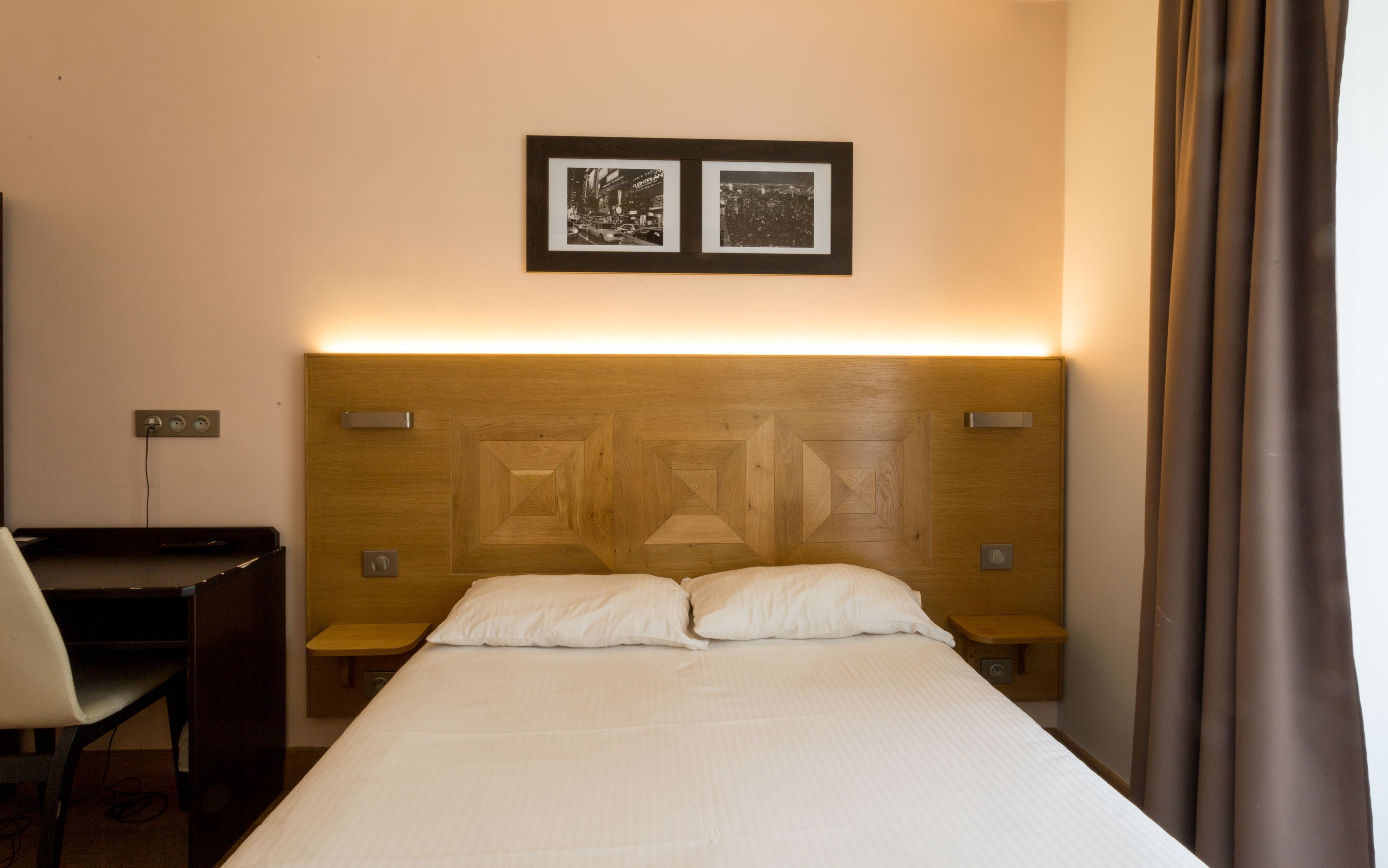 A hotel room with a wooden headboard, two bedside tables, a desk, and a chair. There are two framed pictures on the wall above the bed.