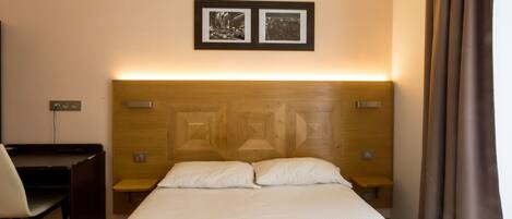 A hotel room with a wooden headboard, two bedside tables, a desk, and a chair. There are two framed pictures on the wall above the bed.