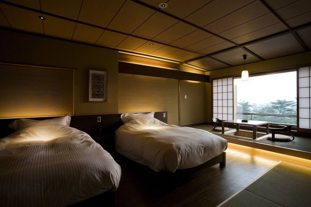 Een hotelkamer met twee bedden, een bureau en uitzicht op bomen door grote ramen.