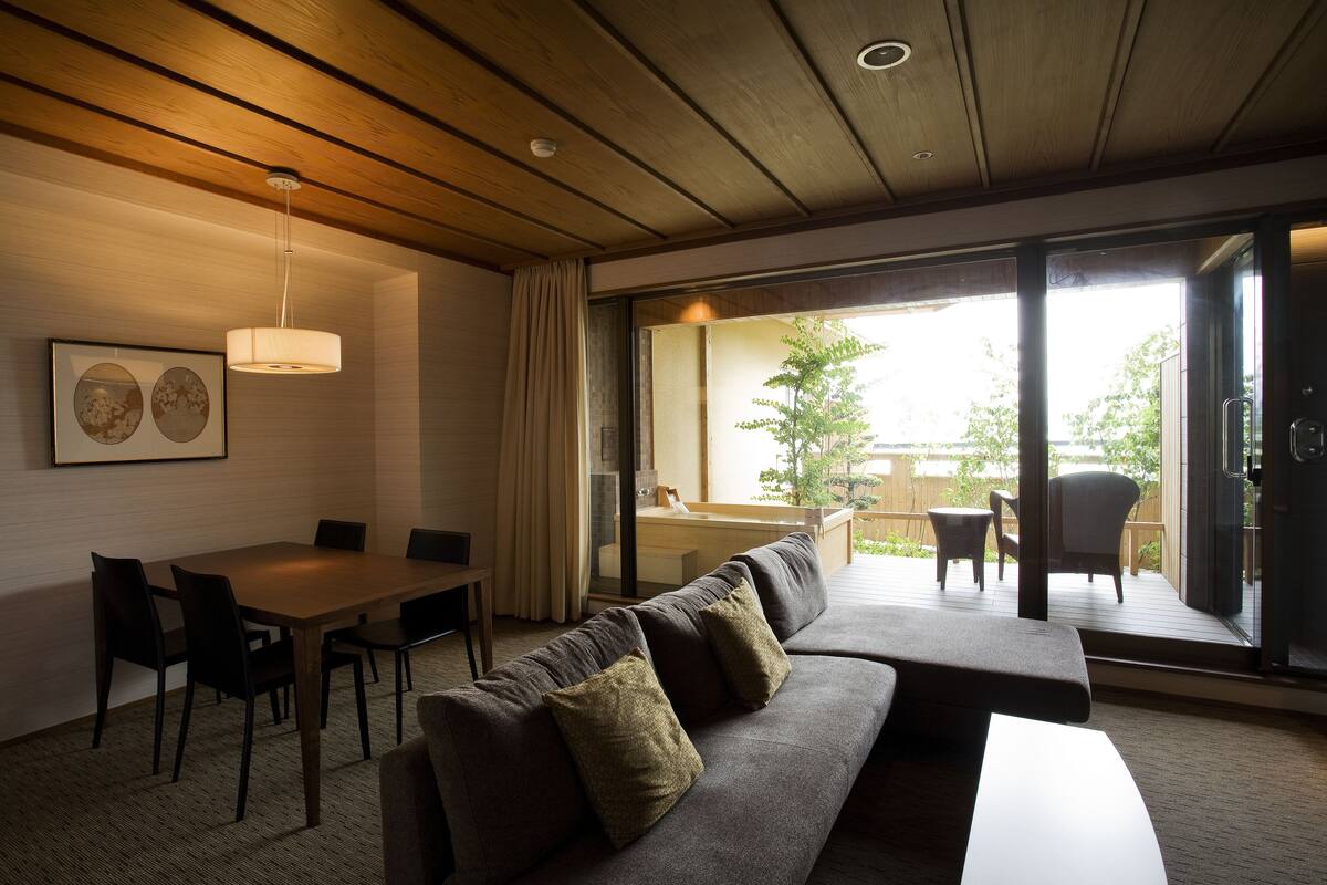 Een moderne woonkamer met een houten plafond, een bank met kussens, een eettafel met stoelen en een grote schuifdeur die leidt naar een buitengebied met een terras en stoelen.