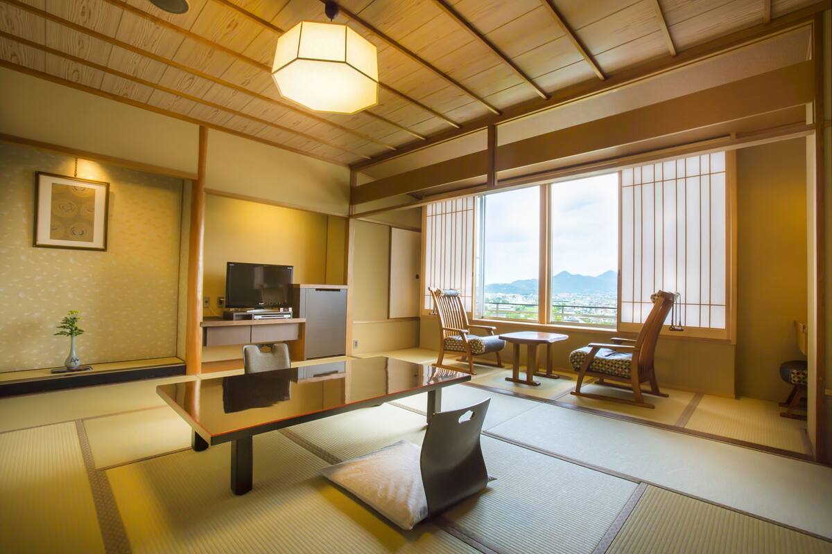 Een traditionele Japanse kamer met tatami vloeren, een lage tafel, een televisie en uitzicht op bergen door grote ramen.