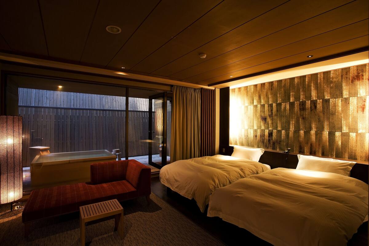 Een moderne hotelkamer met een groot bed, een bank en een kleine tafel. Er is een groot raam met gordijnen en uitzicht op de omgeving.