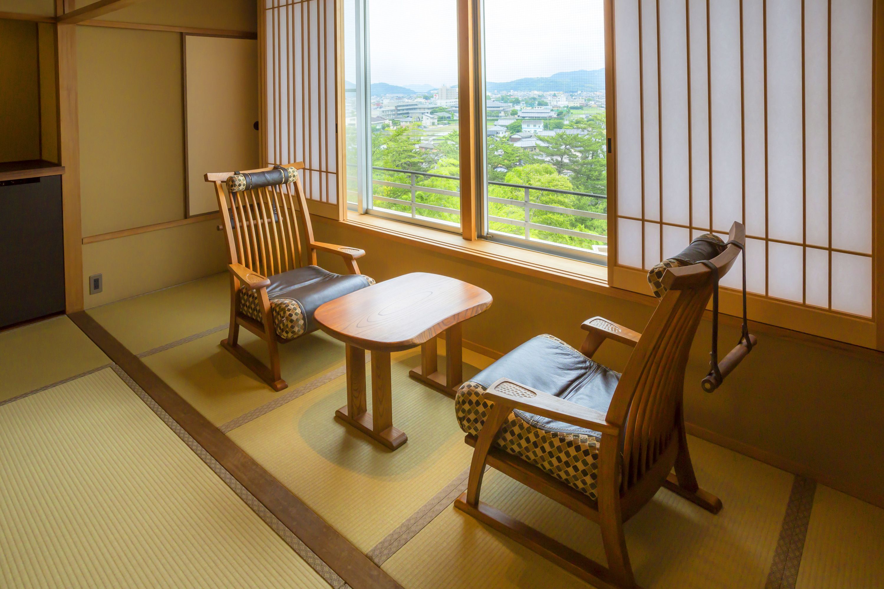 A traditional Japanese room with tatami flooring, a low table, two wooden chairs, and a view of a cityscape through shoji screens.