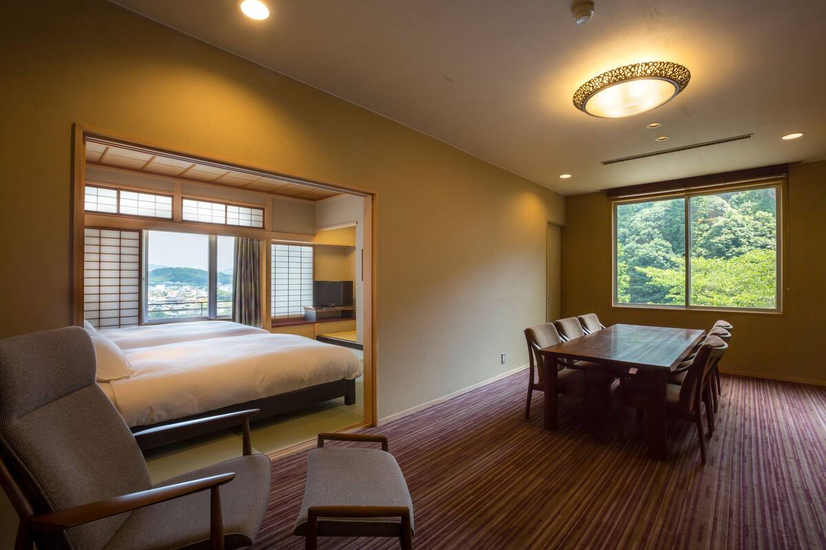 Hotelkamer met een bed, een eettafel met stoelen, een bank en uitzicht op groen via grote ramen.