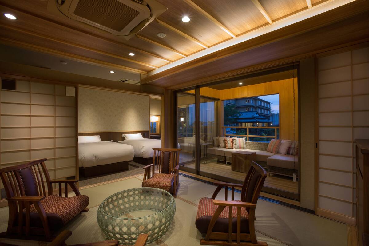 Een hotelkamer met een traditioneel Japans interieur, met tatami vloeren, een laag houten plafond en schuiframen naar een balkon met uitzicht op de stad.