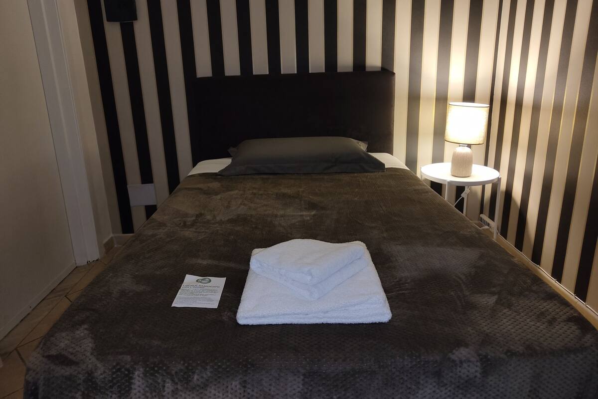 A neatly made bed with a folded white towel, a bedside lamp, and a small card on the floor.