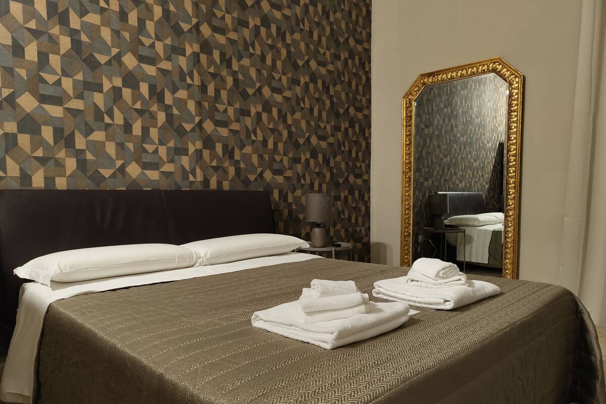 A hotel room with a geometric patterned wall, a large mirror with a gold frame, a neatly made bed with white linens, and a small bedside table with a lamp.