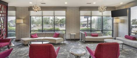 A modern lounge area with sofas, chairs, and a coffee table. Large windows provide a view of the outdoors.