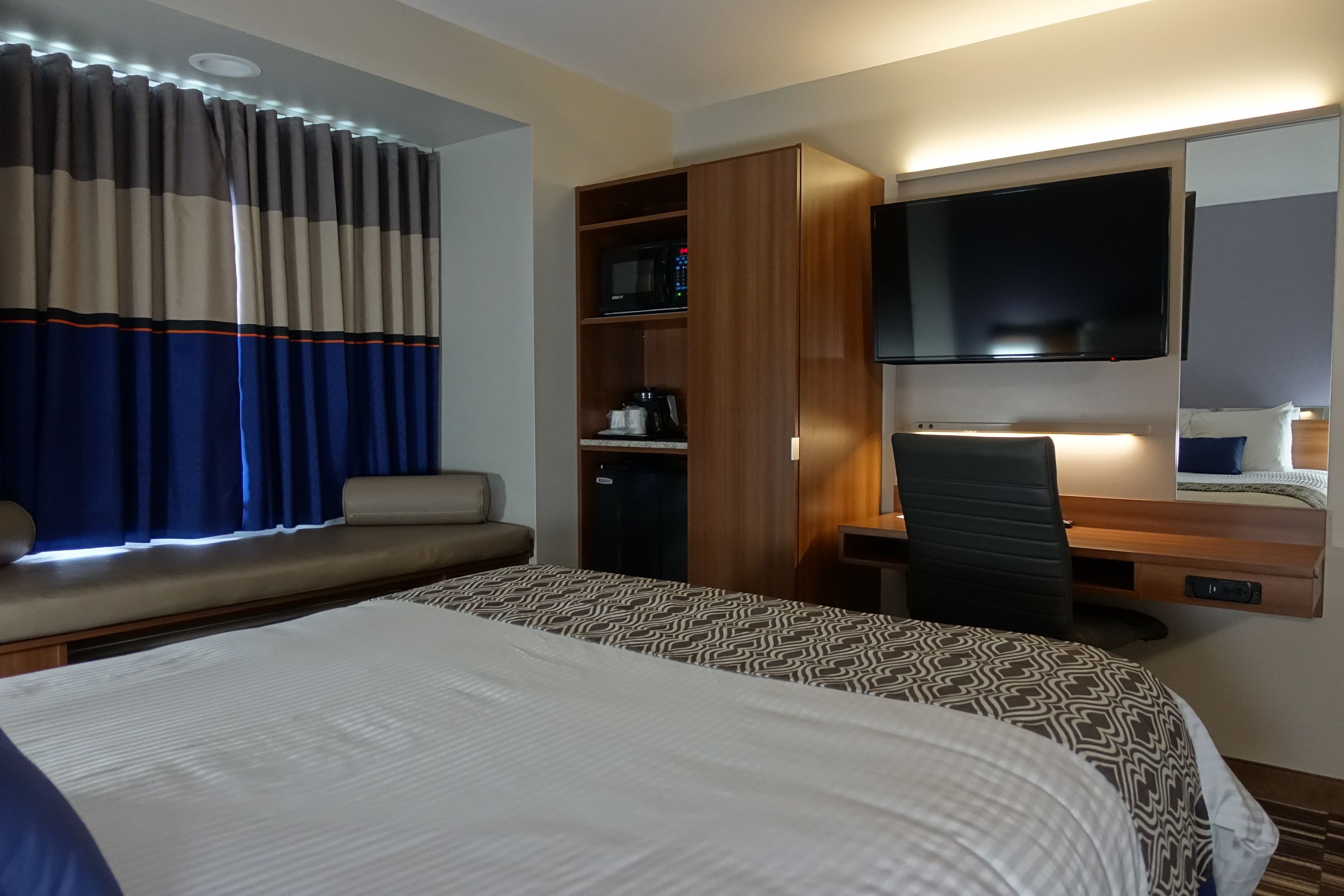 Habitación de hotel con una cama grande, un escritorio con silla, televisor de pantalla plana, minibar y una zona de estar con cortinas.