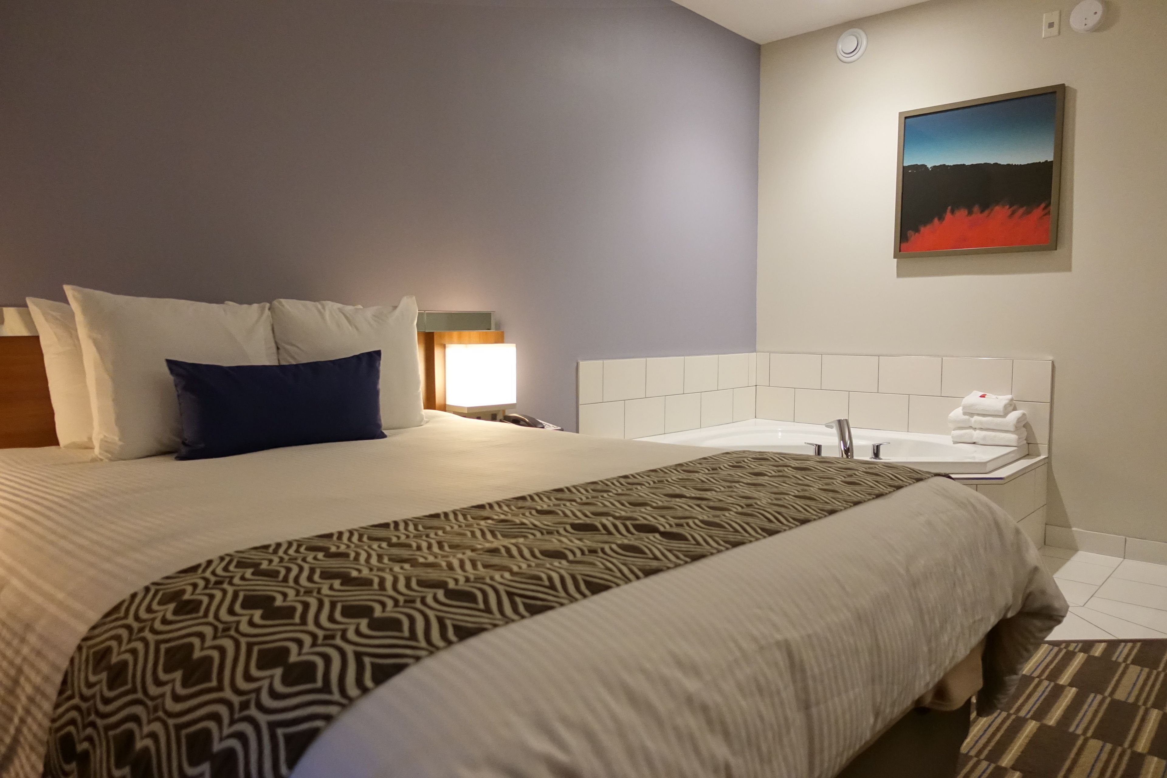 Una habitación de hotel con una cama grande, una mesita de noche, una lámpara, un cuadro en la pared y un baño con lavamanos y bañera.