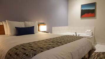 Una habitación de hotel con una cama grande, una mesita de noche, una lámpara, un cuadro en la pared y un baño con lavamanos y bañera.