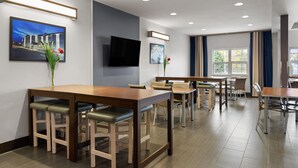 A modern hotel dining area with multiple tables and chairs, a flat-screen TV mounted on the wall, and a large window with curtains. - Microtel Inn & Suites by Wyndham Sault Ste. Marie (Sault Ste. Marie)