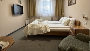 A hotel room with a bed, a television, a window with curtains, a radiator, and a small table with a chair. - Hotel Pansion Stari Grad - Sarajevo (Sarajevo)