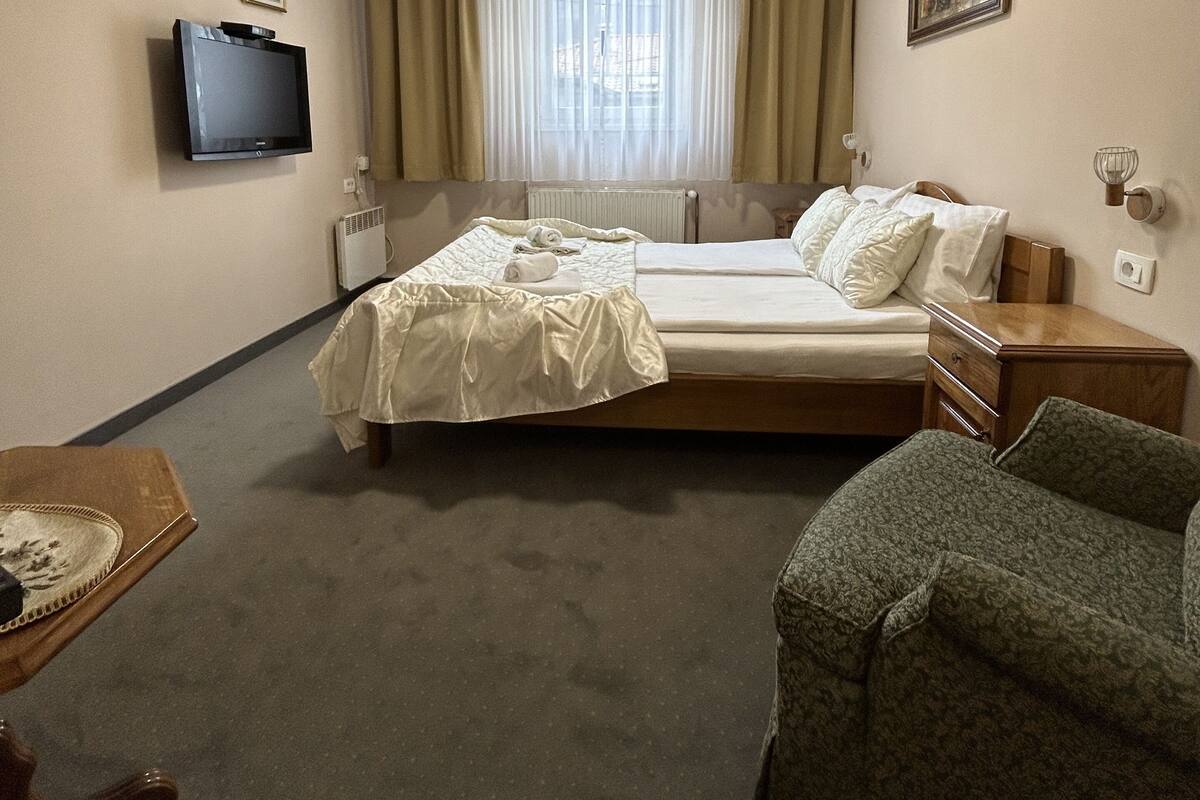 A hotel room with a bed, a television, a window with curtains, a radiator, and a small table with a chair.