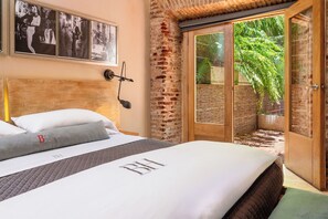 A bedroom with a large bed, a headboard, a wall-mounted lamp, and a sliding glass door leading to an outdoor area with a brick wall and greenery. - Billini Hotel, Historic Luxury (Santo Domingo)