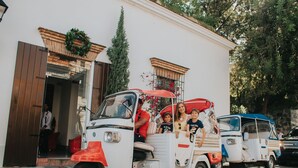 A white and red vintage vehicle with passengers, parked in front of a building with a wooden door and a wreath on the wall. - Billini Hotel, Historic Luxury (Santo Domingo)