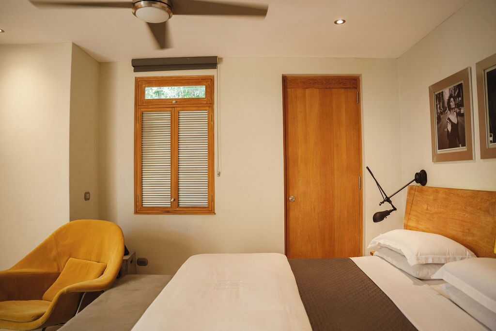 A bedroom with a bed, a chair, a window with blinds, a door, and a ceiling fan.