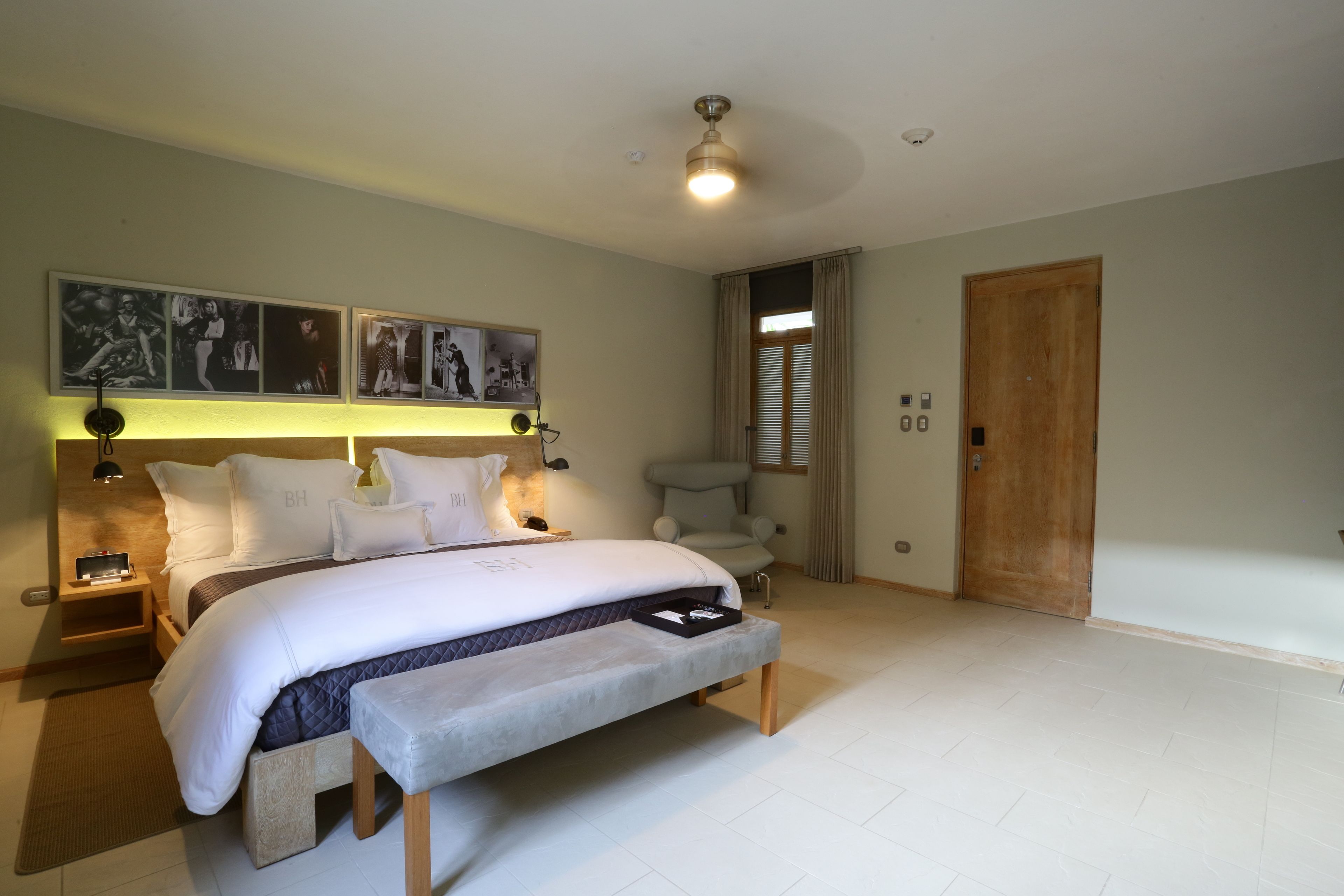 A bedroom with a large bed, bedside tables, a bench, a ceiling fan, and a door.