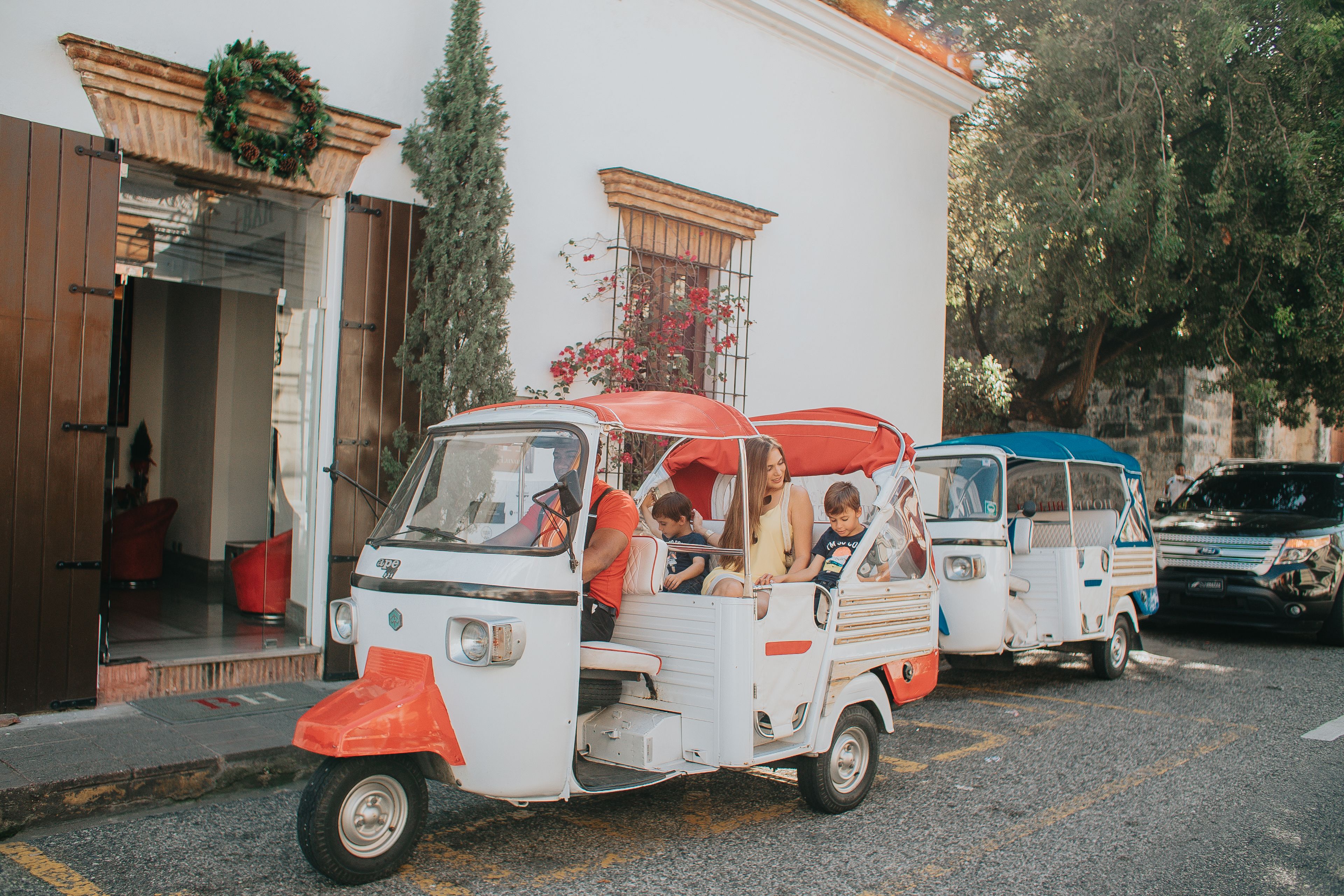 Um tuk-tuk branco e vermelho com passageiros, estacionado na rua em frente a um prédio com porta de madeira e janela com grinalda.