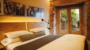 A hotel room with a large bed, a wooden headboard, a nightstand, a desk lamp, and a window with a view of greenery. - Billini Hotel, Historic Luxury (Santo Domingo)