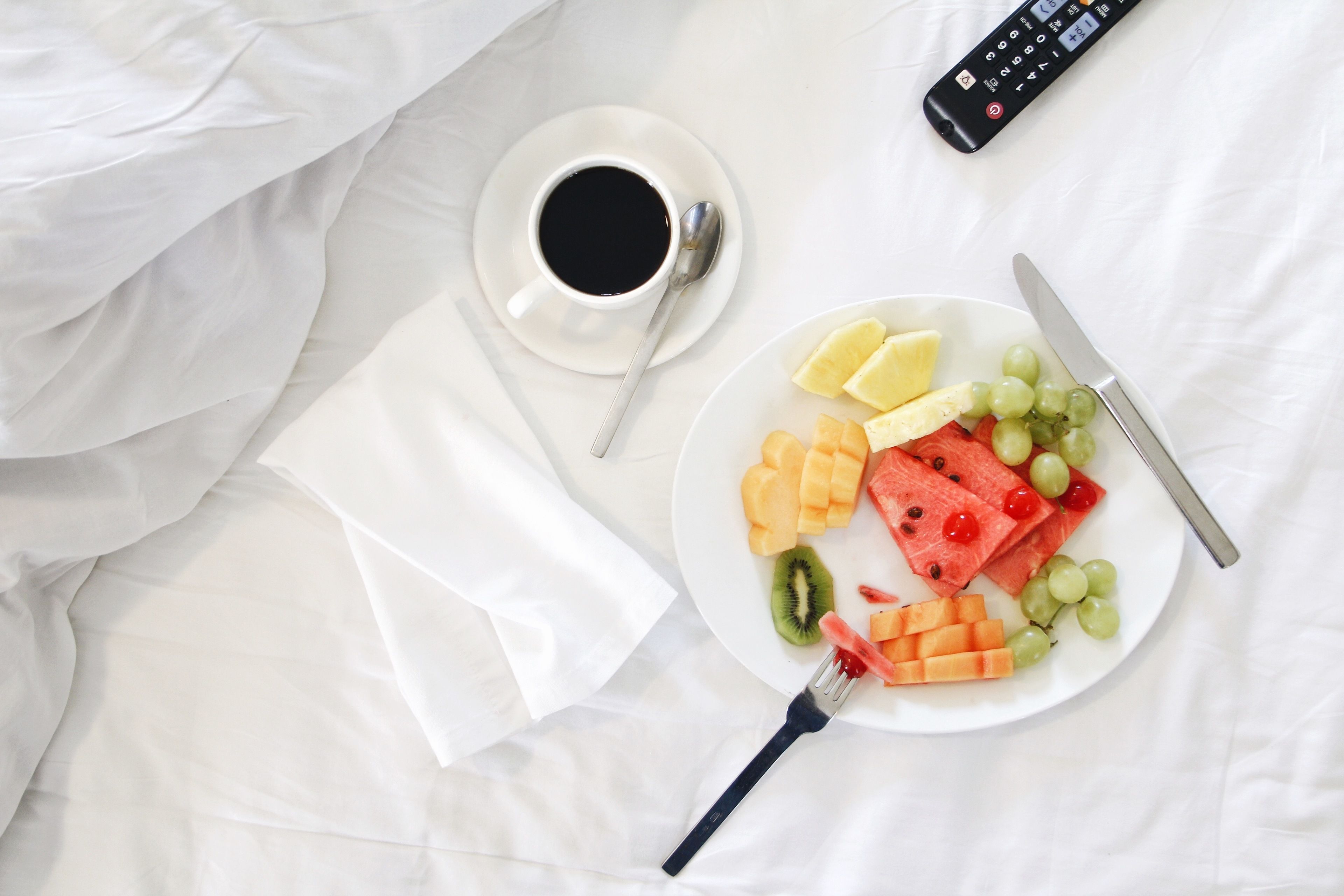 Una coperta bianca con un piatto di frutta, una tazza di caffè, una forchetta, un coltello e un telecomando.