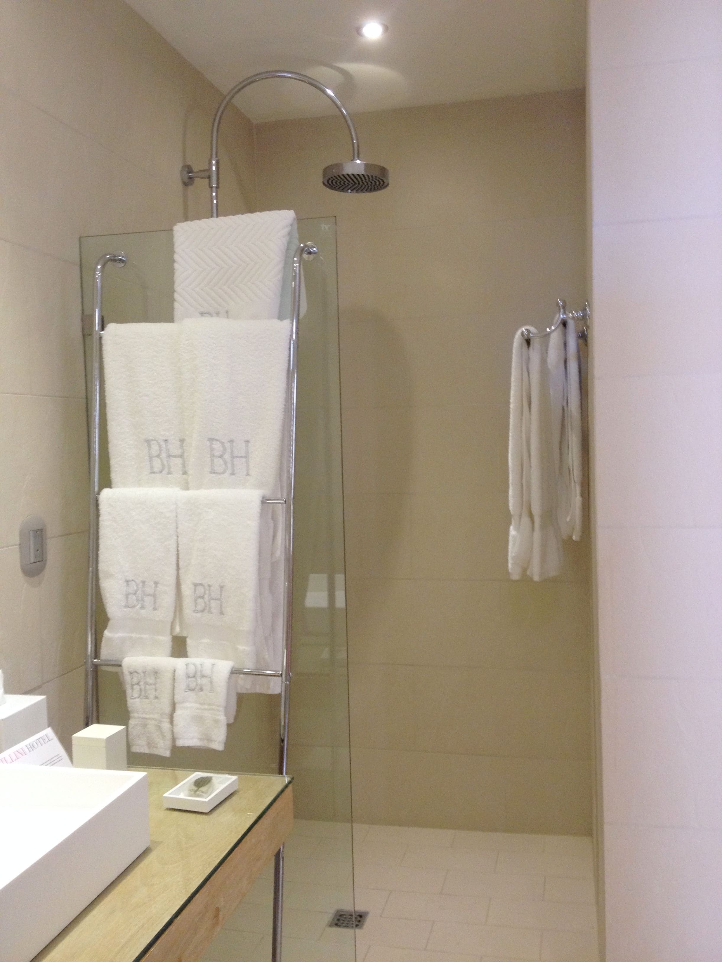 A modern bathroom with a glass-enclosed shower, a white sink, and multiple white towels with "BH" monogrammed on them.