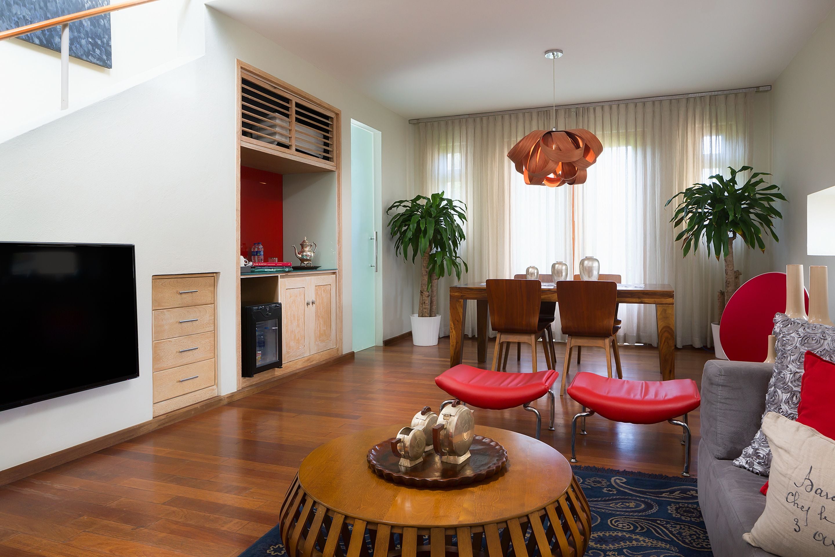 A modern living room with a flat-screen TV, wooden coffee table, red cushioned seating, and a dining area with a wooden table and chairs.
