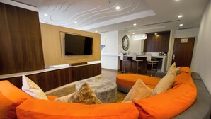 Een moderne woonkamer met een gebogen grijze bank, oranje kussens, een flatscreen televisie en een keuken in de achtergrond.