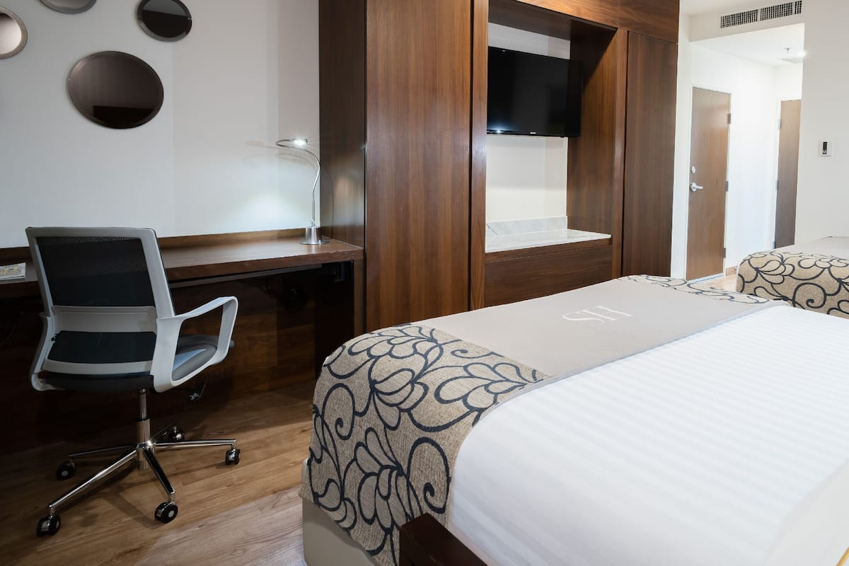 Habitación de hotel con dos camas, un escritorio con silla, un televisor y un cuadro en la pared.