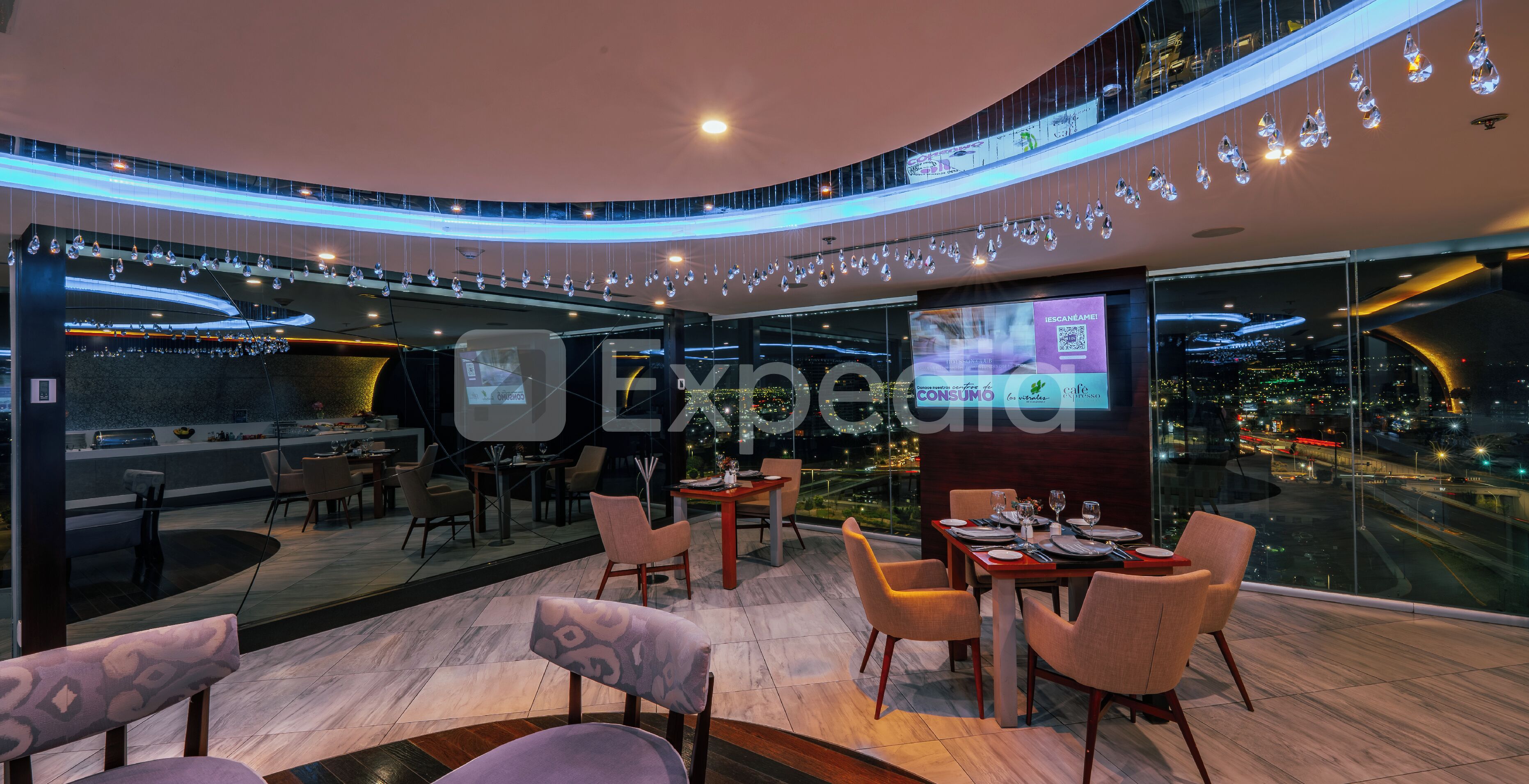 A modern restaurant with a panoramic city view, a digital screen, and a ceiling adorned with hanging crystal lights.