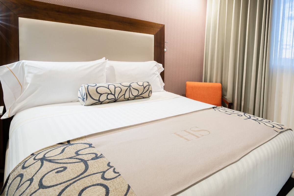 Habitación de hotel con cama, cabecera, mesita de noche, una silla naranja y una ventana con cortinas.
