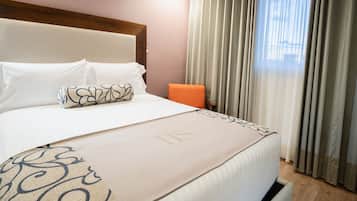Habitación de hotel con cama, cabecera, mesita de noche, una silla naranja y una ventana con cortinas.