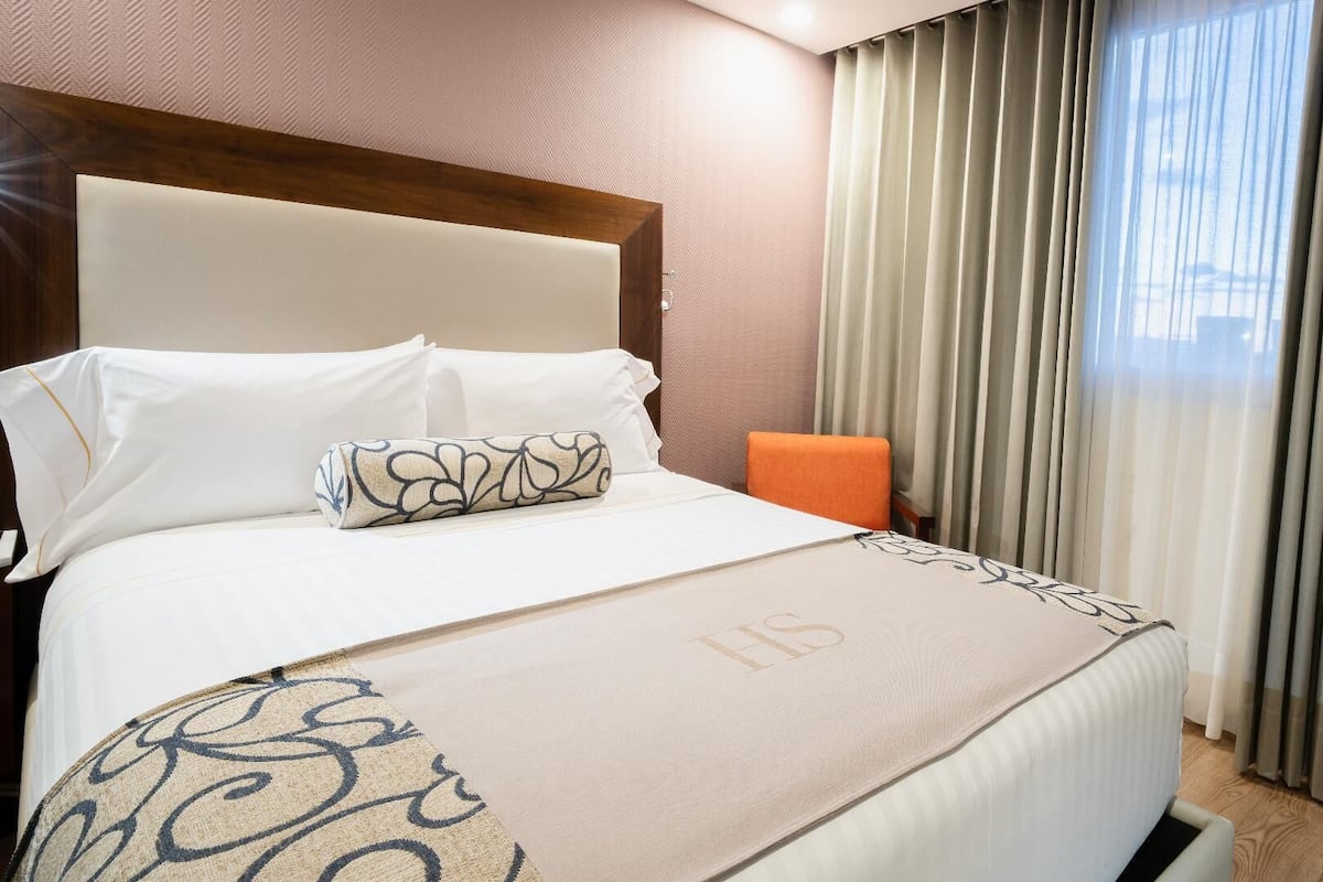 Habitación de hotel con una cama grande, cabecera, mesita de noche, una silla naranja y una ventana con cortinas.