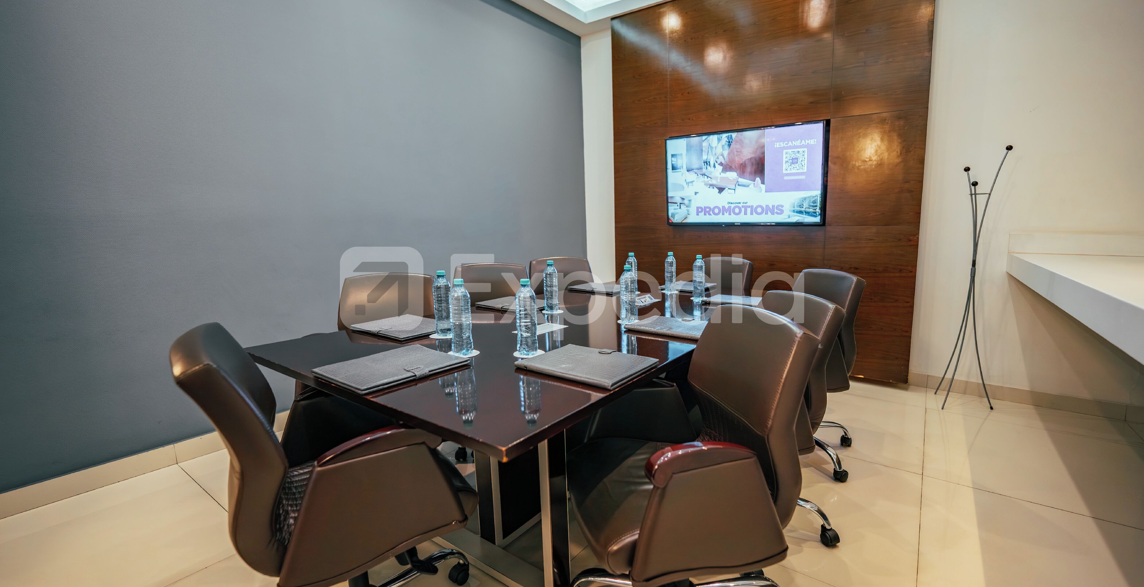 Una sala de conferencias con una pantalla grande mostrando una presentación, botellas de agua sobre la mesa y una pared de acento de madera.