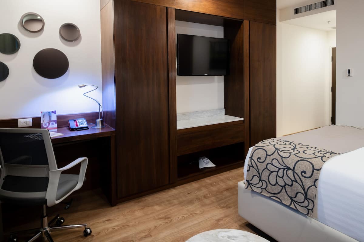Un dormitorio con una cama, un escritorio con silla, un televisor montado en la pared y un cuadro en la pared.