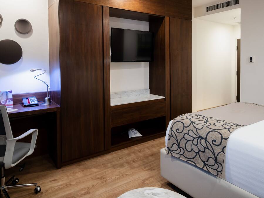 Un dormitorio con una cama, un escritorio con silla, un televisor montado en la pared y un cuadro en la pared.