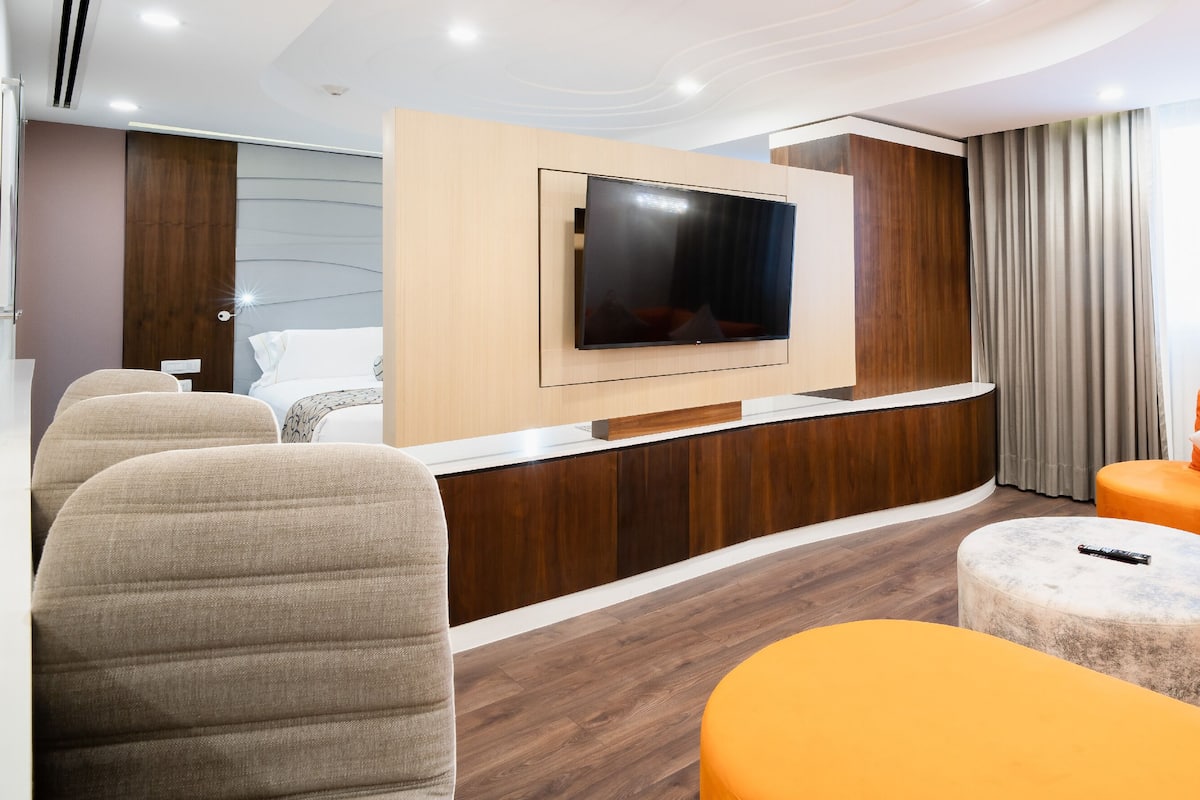 Una habitación de hotel moderna con un televisor de pantalla plana montado en un panel de madera, un sofá con cojines anaranjados y un otoman redonda.