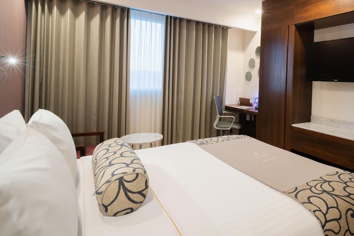 Habitación de hotel con una cama grande, un escritorio con silla, televisor montado en la pared y una ventana con cortinas.