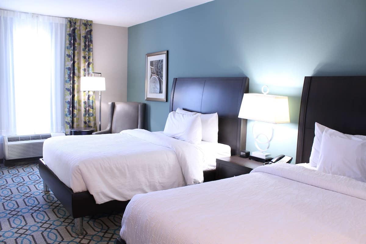 A hotel room with two beds, a desk, a chair, a lamp, and a framed picture on the wall.