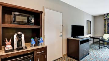 A hotel room with a kitchenette including a microwave, coffee maker, and dishwasher. There is a television, a desk, and a chair.