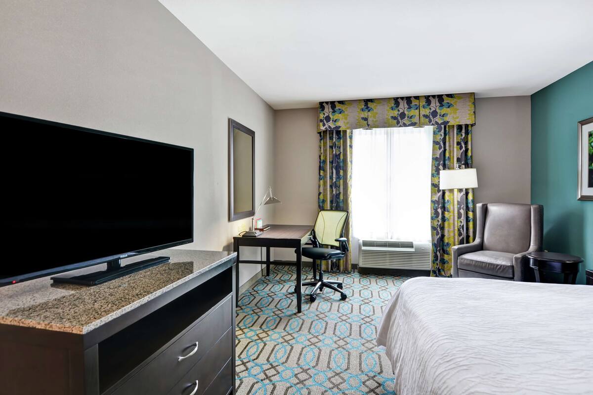 A hotel room with a flat-screen TV, a desk with a chair, a bed, and a window with curtains.