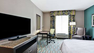 A hotel room with a flat-screen TV, a desk with a chair, a bed, and a window with curtains.