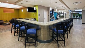 Un espace bar moderne doté d’un comptoir en granit aux lignes courbes, de tabourets de bar noirs et d’un téléviseur à écran plat fixé au mur.