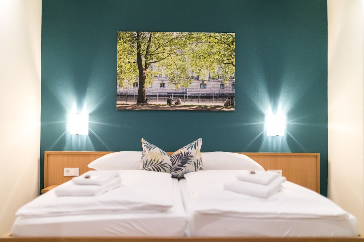 A neatly made bed with white linens and a pillow with a leaf pattern, a wall-mounted picture of a tree, and two wall-mounted lights.