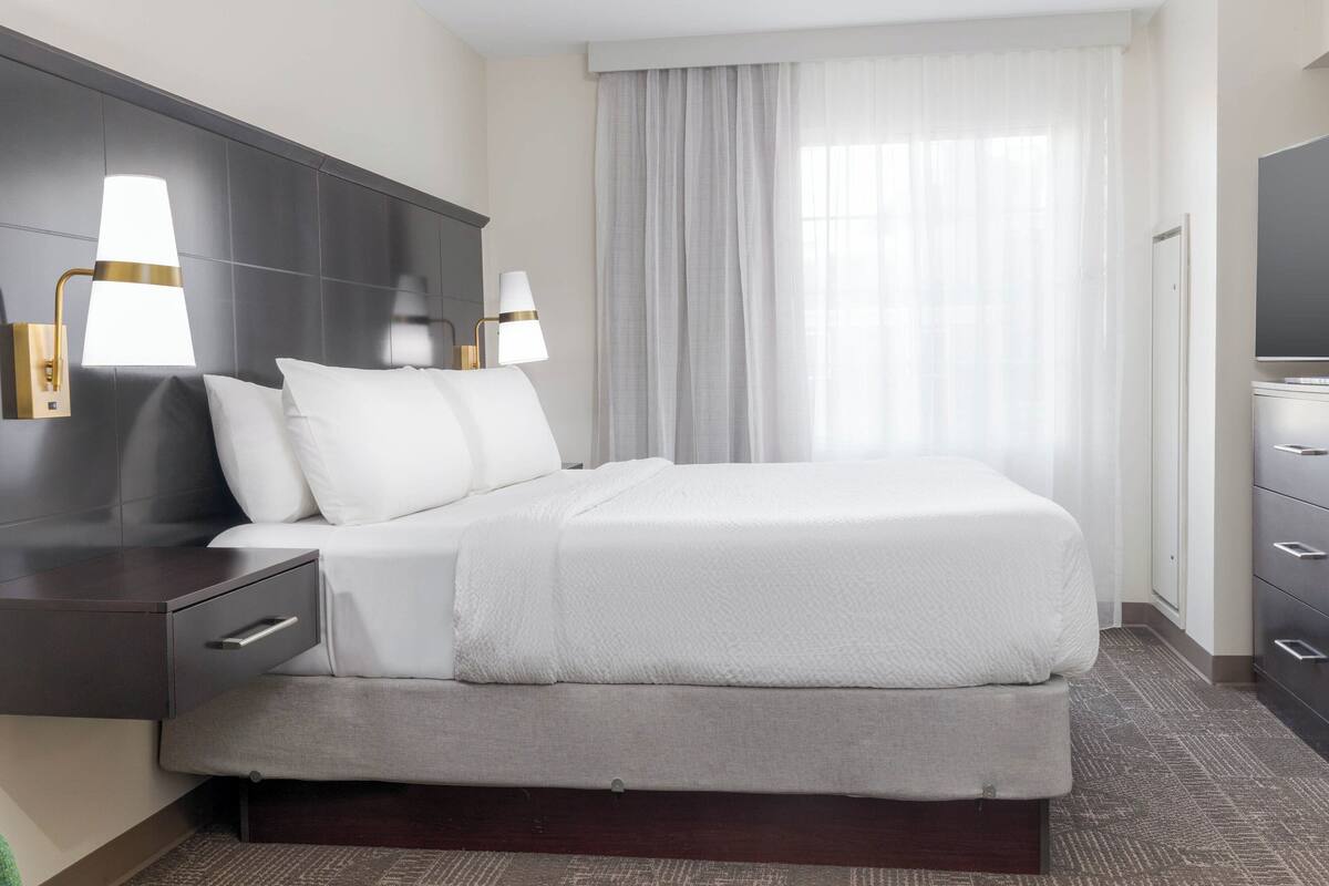 Une chambre d’hôtel moderne dotée d’un grand lit, de deux tables de chevet avec des lampes, d’une tête de lit sombre, d’une grande fenêtre avec des rideaux légers et d’un téléviseur à écran plat fixé au mur.