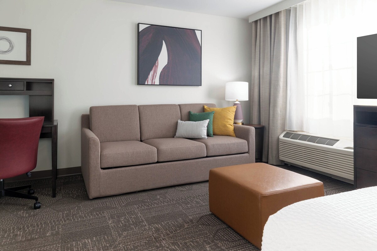 A modern hotel room with a sofa, a red chair, a brown ottoman, a desk with a lamp, and a large window with curtains.