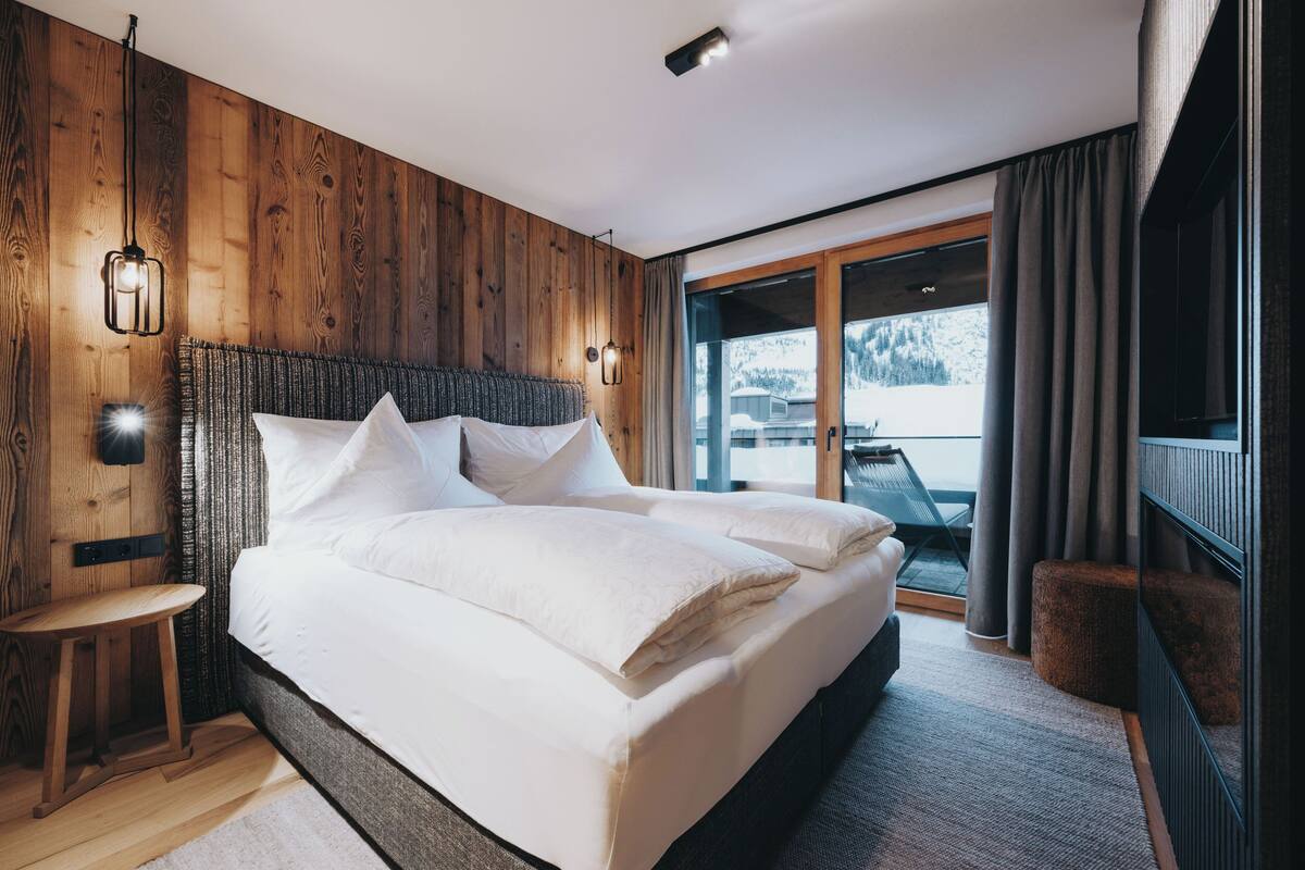 A modern bedroom with a wooden wall, a large bed, a small table, and a view of the outdoors through a sliding door.