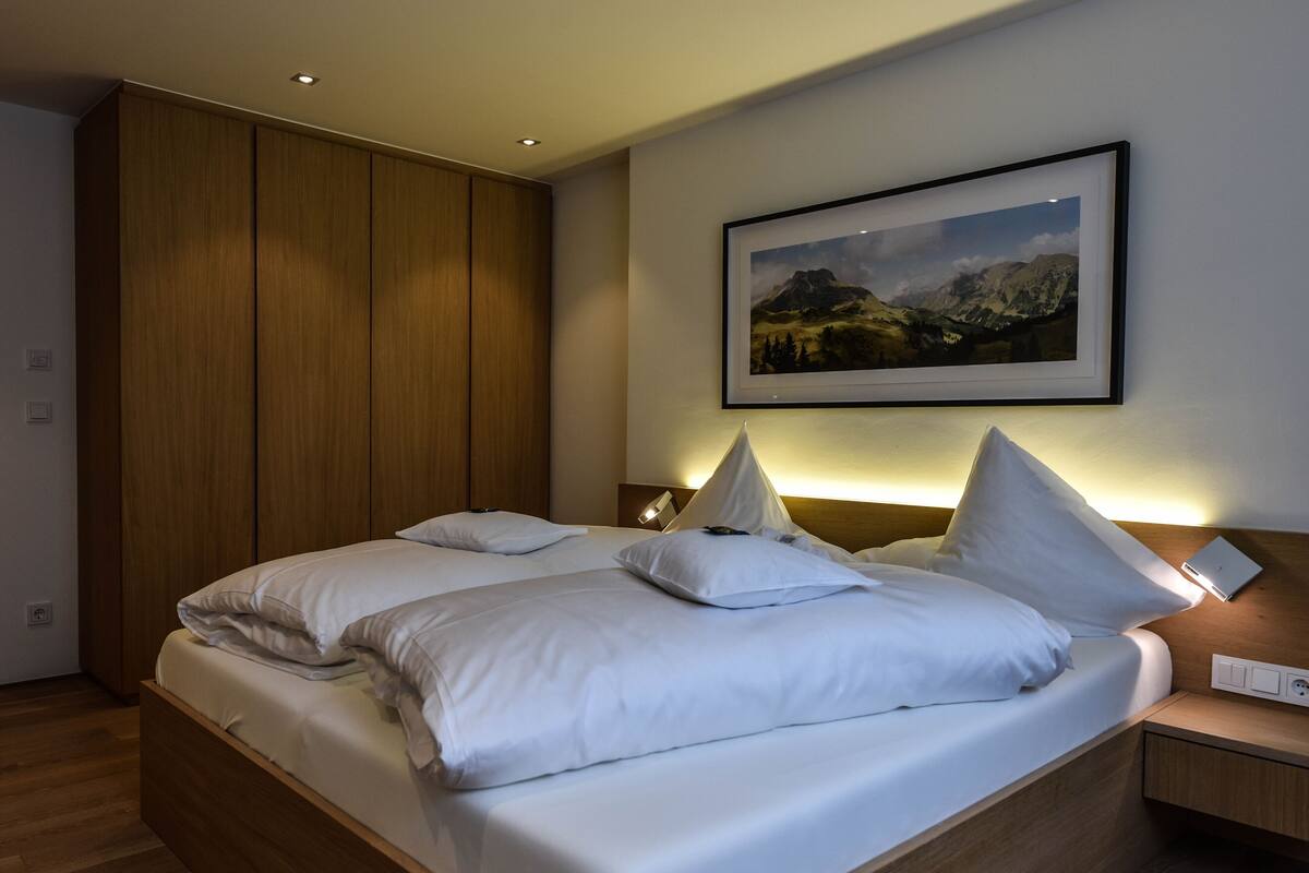 A neatly made double bed with white linens, a wooden headboard, and a framed landscape painting above it.