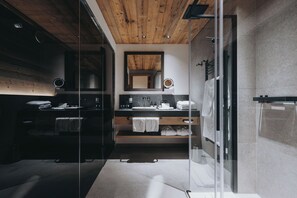A modern bathroom with a wooden ceiling, a glass shower enclosure, a vanity with a mirror, and a bed with towels. - Balma Restaurant | Suites (Lech am Arlberg)